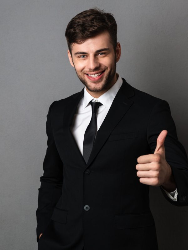 Young successful businessman in formal wear showing thumb up gesture, looking at camera, isolated on gray background